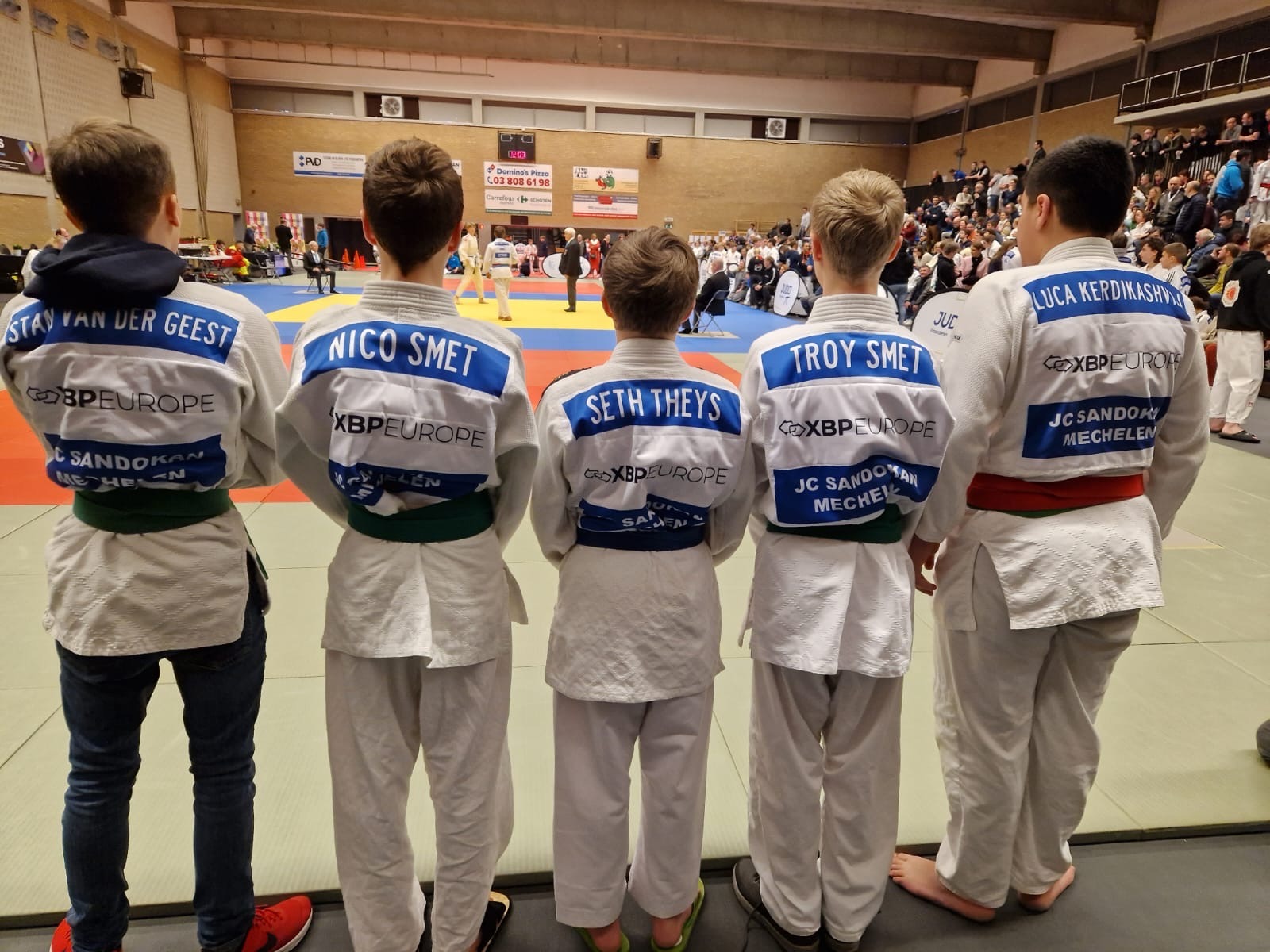 Clubleden van Judoclub Sandokan Mechelen staan met hun rug naar de camera terwijl ze een wedstrijd aanschouwen