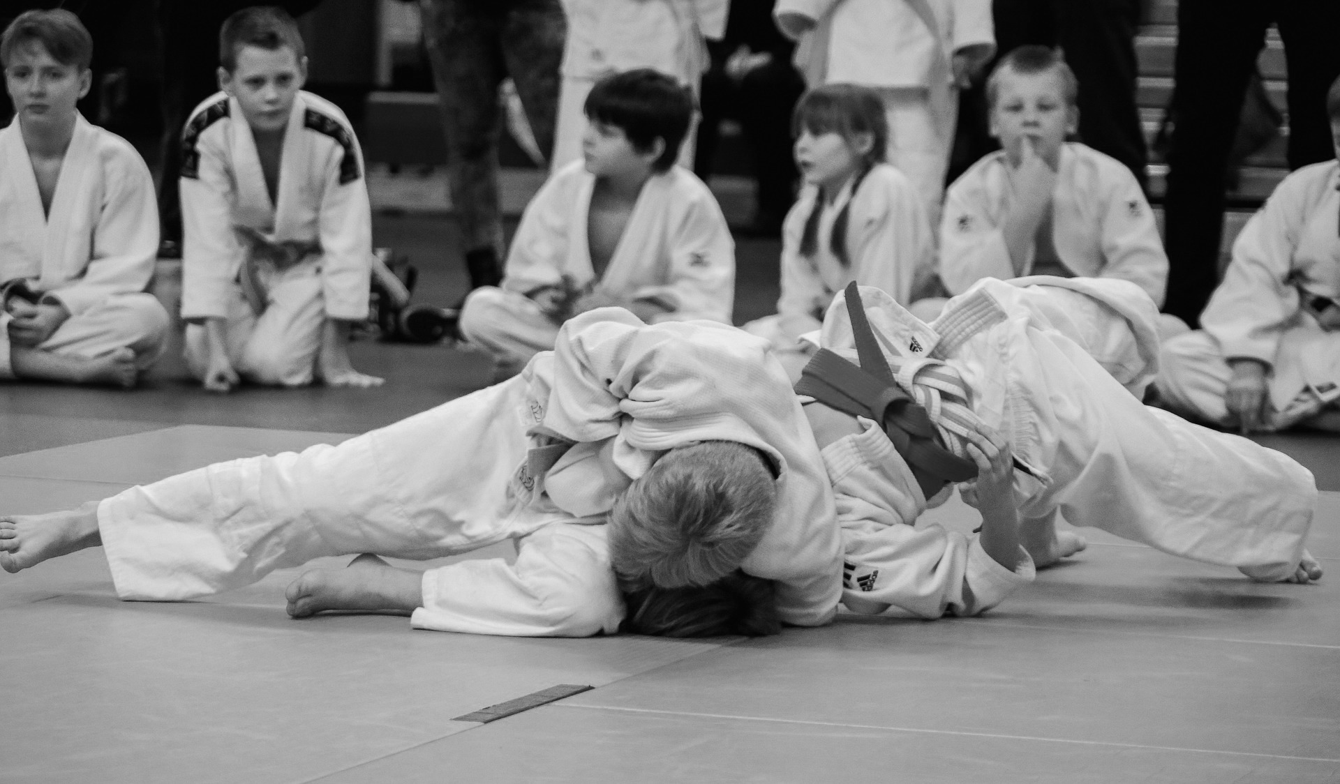 Twee jonge judoka's voeren een judo grondtechniek uit, terwijl op de achtergrond een aantal andere kinderen aan de zijlijn zitten om toe te kijken