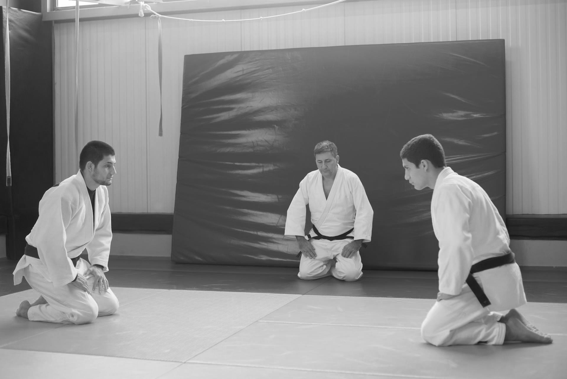 Twee judoka's zitten op de knieën tegenover elkaar op een trainingsmat en groeten elkaar, terwijl hun coach toekijkt al knielend aan de zijkant