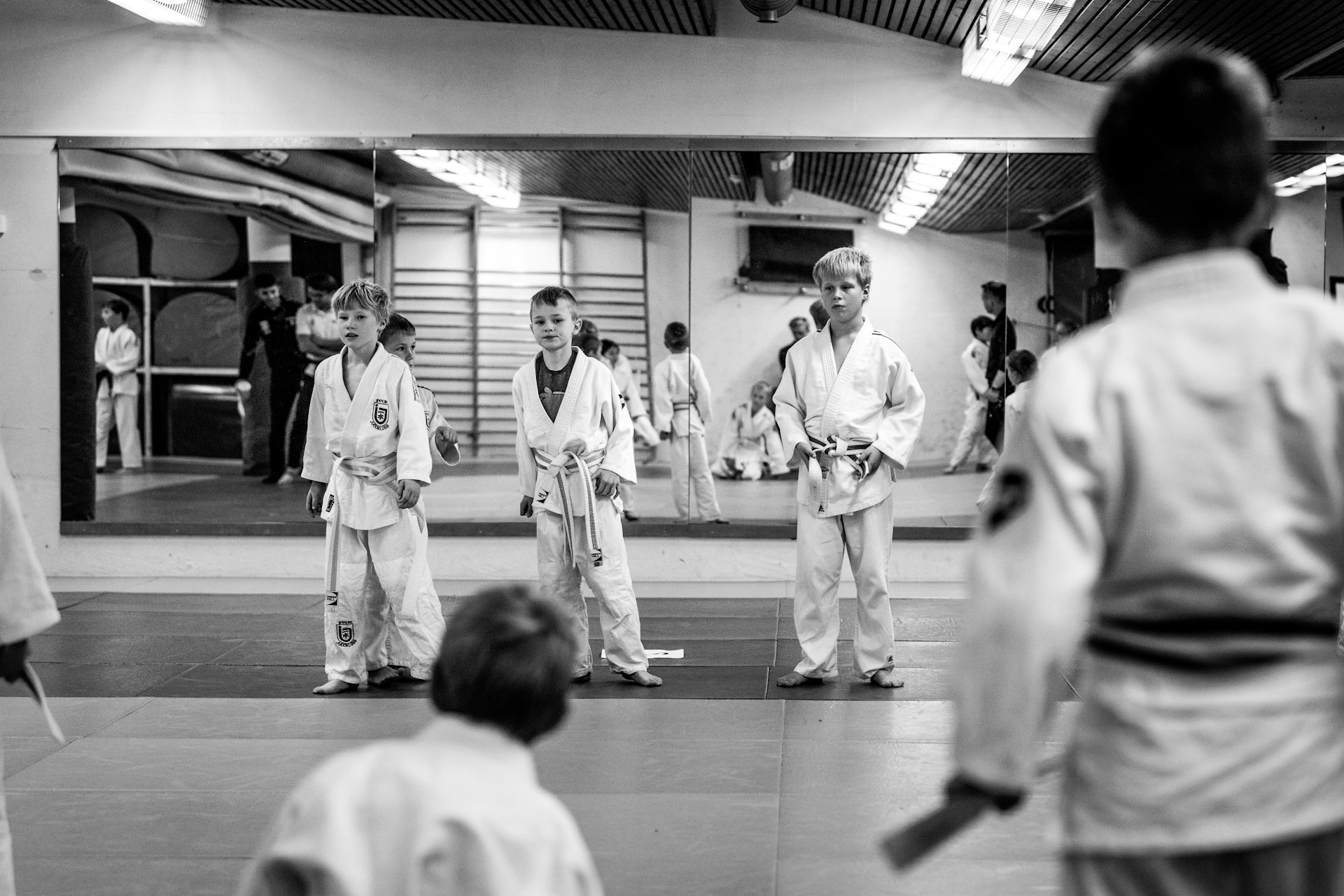 Kinderen in judogi staan op een rij op een trainingsmat in een sportzaal met een spiegelmuur, waarin andere jonge judo beoefenaars te zien zijn